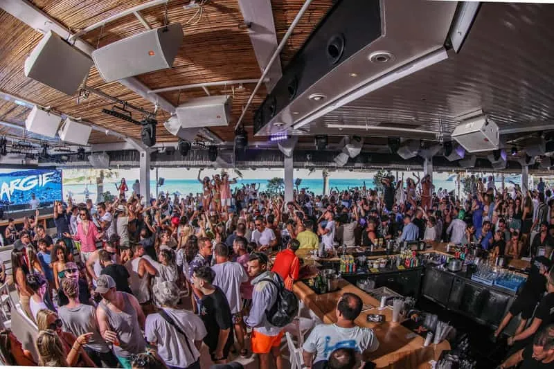 Crowds dancing at a Mykonos beach club day party with DJs and Aegean Sea backdrop during Mykonos island nightlife